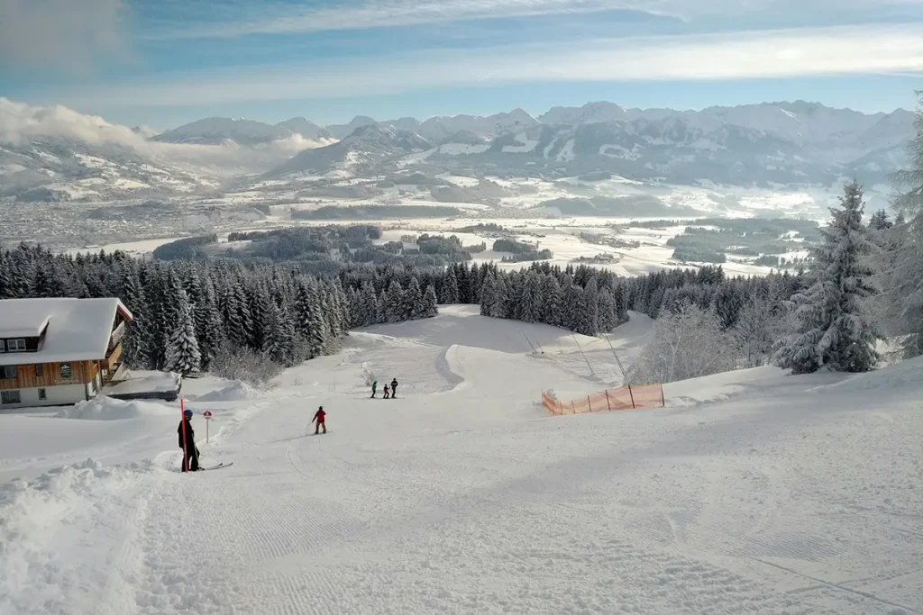 Grande piste de ski animée, vacanciers en descente entourés de montagnes et forêts enneigées